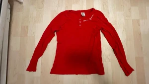 Röd långärmad topp med broderi - Snygg röd långärmad topp med små hålmönster och vit broderad text vid halsen. Toppen har knappar framtill och är tillverkad i ett mjukt material som känns skönt mot huden. Perfekt för dig som vill ha en enkel men ändå unik look.