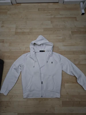 Vit hoodie med dragkedja från Polo Ralph Lauren - Säljer en stilren vit hoodie från Polo Ralph Lauren med dragkedja framtill och två stora fickor. Klassisk huva med dragsko och den ikoniska lilla loggan broderad på bröstet. Perfekt för en clean och avslappnad look. Funkar ferfekt och är äkta, Är M men känns som S. Priset kan diskuterad