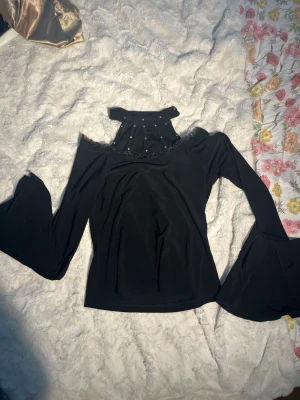 Svart topp med utsvängda ärmar - Snygg svart topp med hög hals och pärldetaljer. Toppen har off shoulder-design och vida, utsvängda ärmar som ger en cool retrovibe. Materialet ser stretchigt och mjukt ut, perfekt för dig som vill sticka ut lite extra. Ärmarna har en slits. Väldigt unik topp som inte längre är min stil. Köpt secondhand och har ingen lapp kvar så vet inte märket.