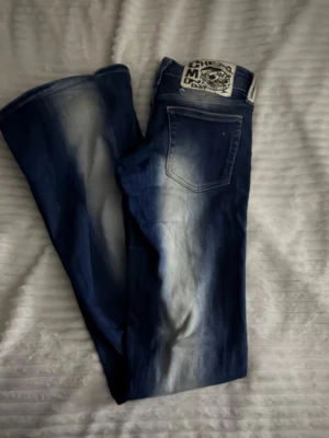 Mörkblå bootcut jeans med slitningar - Säljer ett par mörkblå bootcut jeans med snygga slitningar och ljusa tvättade partier framtill och baktill. Jeansen har klassisk femficksmodell och låg midja. Materialet är denim i bomull. Perfekta för en avslappnad och trendig look.