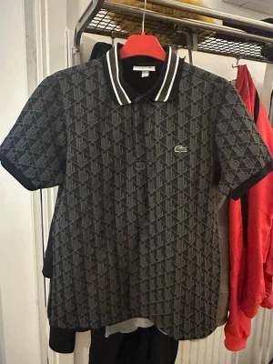 Svart mönstrad piké från Lacoste - Snygg svart pikétröja från Lacoste med diskret geometriskt mönster och klassisk krage med vita ränder. Ikoniska krokodilloggan på bröstet. Perfekt för en stilren och sportig look.