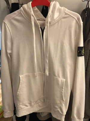 Vit hoodie med dragkedja Stone Island - Säljer en vit hoodie från Stone Island med dragkedja framtill, två stora fickor och klassisk huva med snören. Ikonisk svart logopatch på vänster ärm. Tillverkad i mjukt bomullsmaterial som känns skönt mot huden. Perfekt för en clean och stilren look.