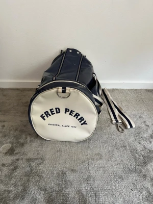 Marinblå duffelväska Fred Perry - Snygg marinblå duffelväska från Fred Perry med vita detaljer och avtagbar axelrem. Väskan har rund form, dragkedja upptill och tydlig logga på sidan. Tillverkad i glansigt konstläder och har både handtag och axelrem för olika bärsätt. Om du har frågar är det bara att fråga på. Pris går att diskutera med seriösa köpare. Kolla in min profil och se om du hittar något mer intressant. Mvh Larsen_Closet
