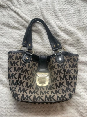 Beige och svart MK handväska - Snygg handväska från Michael Kors i beige canvas med svart MK-logga över hela väskan. Väskan har svarta handtag i läder och en stor guldfärgad låsdetalj framtill. Perfekt för dig som gillar statement-accessoarer.