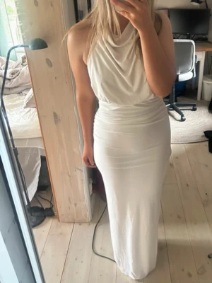 Vit långklänning med draperad hals - Fin vit klänning med öppen rygg, går ner till fötterna på mig som är 1,67. Använd max 5 gånger, har en sminkfläck på kragen vid halsen, inget som syns när man har klänningen på sig eftersom det är på insidan. 