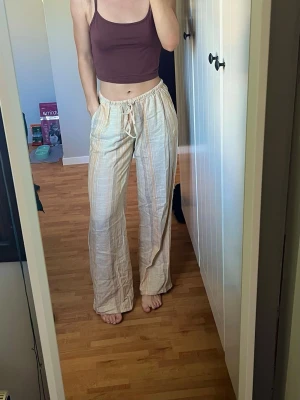 Beige randiga linnebyxor med dragsko - Liinnebyxor i beige med tunna ränder i ljusgult och orange. Byxorna har breda ben, resår i midjan och snöre.