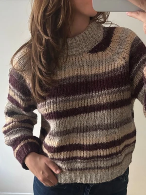 Randig stickad tröja i beige och vinrött - Mysig stickad tröja med breda ränder i beige, grått och vinrött. Tröjan har en hög, ribbad krage och långa ärmar med ribbade muddar. Perfekt för kalla dagar och har en avslappnad, boxig passform. 