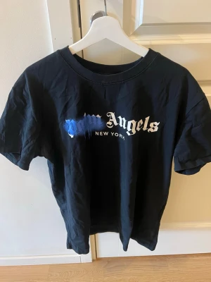 Svart Palm Angels t-shirt New York - Säljer en svart t-shirt från Palm Angels med New York-tryck framtill. T-shirten har korta ärmar och en cool blå färgdetalj över loggan. Klassisk passform och rund halsringning. Perfekt för dig som gillar streetwear och vill sticka ut.