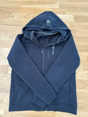Svart hoodie från Sail Racing - Säljer en svart hoodie från Sail Racing med dragkedja framtill och broderad logga på huvan. Hoodien har två sidofickor och mjukt fleecefoder på insidan. Snygga detaljer med vita sömmar och tryck på bröstet. Perfekt för en avslappnad och sportig stil.