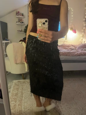 Svart glittrig långkjol med fransar - Säljer en svart långkjol med glittriga detaljer och fransar som ger en riktigt cool look. Kjolen har rak passform och är perfekt för dig som vill sticka ut lite extra. Materialet känns syntetiskt och skimrar snyggt i ljuset. Egentligen high/mid waist men går lätt att få till low waist. 