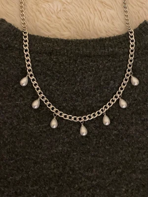 Silverfärgat halsband med pärldroppar - Snyggt halsband i silverfärgad rostfritt med sju droppformade pärlor som hänger längs kedjan. Kedjan är lite grövre och ger ett trendigt intryck. Perfekt för dig som gillar att sticka ut med accessoarer och vill ha något som känns både klassiskt och modernt.
