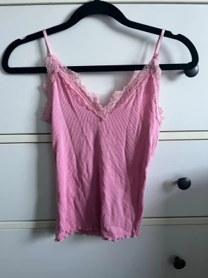 Rosa randig linnetopp med spets - Söt rosa linnetopp med tunna axelband. Toppen har spetsdetaljer vid urringningen och längs kanterna, samt en lätt vågig nederkant. Perfekt för en avslappnad och trendig look.