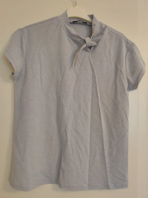 Ljusgrå topp med knyt i halsen - Snygg ljusgrå topp med korta ärmar och unik knytning i halsen. Enkel och stilren design som passar till många olika outfits. Tillverkad i mjukt material för en bekväm känsla.