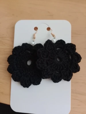 Svarta virkade blomörhängen - Unika örhängen i svart virkat garn formade som blommor. Örhängena har små vita pärlor vid krokarna och metallfästen i guldton. Perfekta för dig som vill sticka ut med en kreativ accessoar.