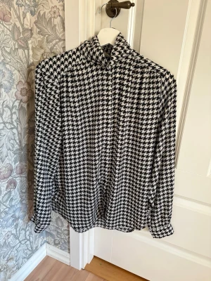 Svartvit rutig skjorta från H&M - Säljer en svartvit skjorta från H&M med klassiskt hundtandsmönster. Skjortan har lång ärm, krage och knäppning framtill. Perfekt för dig som gillar tidlös och trendig stil.