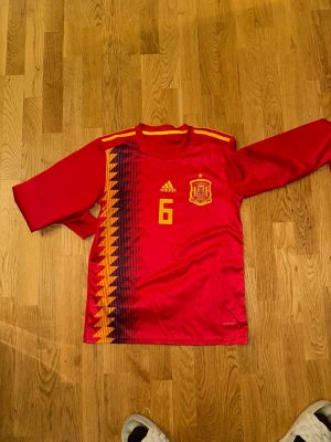Spaniens landslagströja Iniesta #6 - Röd långärmad fotbollströja från Adidas, officiell Spanien landslagströja med nummer 6 och Iniesta på ryggen. Gula detaljer och mönster längs ena sidan, samt landslagets emblem på bröstet. 