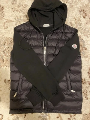 Svart dunväst med huva  - 160-170cm size L i barn size använt 3 ggr bara jätte fin. Snygg svart dunväst med huva och dragkedja framtill. Västen har svarta ärmar i sweatshirtmaterial och en klassisk logga på ena ärmen. Perfekt för lager-på-lager och streetwear-vibe.