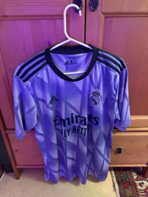 Lila Real Madrid matchtröja Benzema - Säljer en lila Real Madrid matchtröja från Adidas med svart krage och svarta ränder på axlarna. Tryck med Benzema och nummer 9 på ryggen samt La Liga-märke på ärmen. Framsidan har klubbmärke och sponsortryck. Perfekt för fotbollsfans!