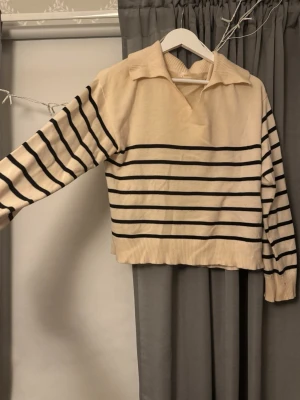 Beige tröja med svarta ränder - Trendig beige tröja med svarta ränder och krage. Tröjan har långa ärmar, ribbade muddar och en avslappnad passform. Perfekt för dig som gillar en klassisk men ändå modern look.