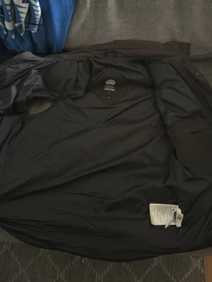 Svart dunväst från Canada Goose - Snygg svart dunväst från Canada Goose med klassisk logga på bröstet. Västen har hög krage, tryckknappar och två fickor framtill. Perfekt för lager-på-lager och håller dig varm under kyliga dagar.