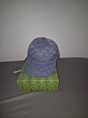 Blå denimkeps från Gucci - Säljer en snygg blå keps från Gucci i denim med det klassiska GG-mönstret över hela kepsen. Kepsen har böjd skärm och justerbar rem bak. Perfekt för dig som gillar exklusiva accessoarer och streetwear. Priset kan diskuteras 