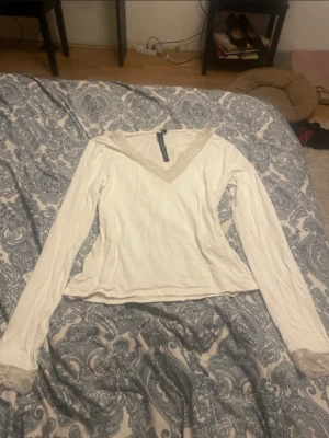 Vit långärmad topp med spetsdetaljer - Säljer en vit långärmad topp i storlek M med v-ringning och fina spetsdetaljer vid halsringningen och ärmsluten. Perfekt att styla med jeans eller kjol för en trendig look.