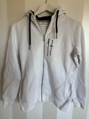 Vit hoodie från Sail Racing - Säljer en vit hoodie från Sail Racing med dragkedja, svart snörning i huvan och en bröstficka med logga. Hoodien har två sidofickor och är tillverkad i mjukt bomullsmaterial. Perfekt för en avslappnad och sportig stil.