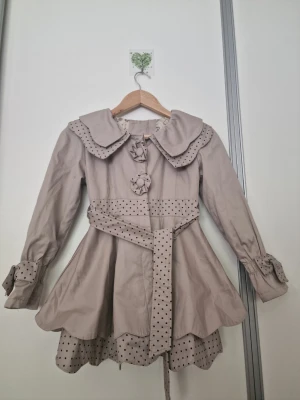 Beige kappa med prickiga detaljer Barn 8-9 år - Supersöt beige kappa med volangkrage, knytband i midjan och dekorativa blommor framtill. Prickiga detaljer på krage, ärmslut och nederkant ger en lekfull touch. Kappan har utsvängd passform och långa ärmar. Perfekt för dig som gillar en gullig och unik stil.
