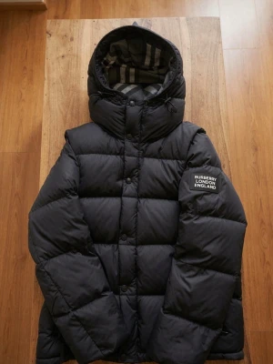 Svart pufferjacka från Burberry - Snygg svart pufferjacka från Burberry med avtagbara ärmar så den kan bäras som väst. Jackan har huva med klassiskt rutigt foder och logotyp på ärmen. Perfekt för kalla vinterdagar och har en modern, boxig passform.