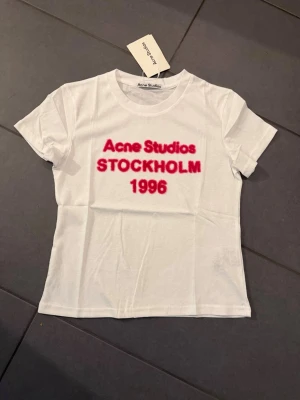Vit Acne Studios t-shirt med tryck M - Vit t-shirt från Acne Studios med rött tryck på bröstet där det står 'Acne Studios STOCKHOLM 1996'. Klassisk rund hals och korta ärmar. Perfekt för en trendig och avslappnad stil.