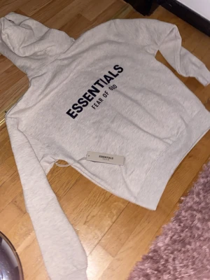 Essentials Fear of God hoodie beige - Snygg beige hoodie från Essentials Fear of God med svart tryck på bröstet. Klassisk passform med huva och ribbade muddar. Tillverkad i mjukt bomullsmaterial som känns skönt mot huden. Perfekt för en avslappnad och trendig look.