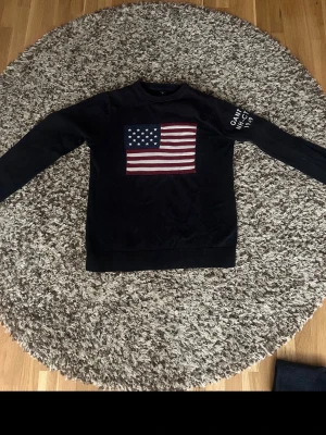 Svart stickad tröja med USA-flagga - Snygg svart stickad tröja med en stor amerikansk flagga på bröstet och vit text på ena ärmen. Tröjan har rund halsringning och långa ärmar. Perfekt för dig som vill ha en statement-piece i garderoben.
