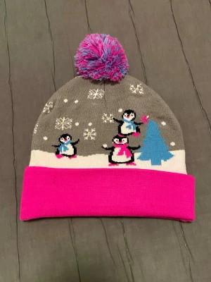 Nya grå mössa med pingviner och tofs - Säljer en grå mössa med rosa uppvik och färgglad tofs. Mössan har ett vintermönster med snöflingor, pingviner och en blå gran. Perfekt för kalla dagar och ger en lekfull touch till din outfit.