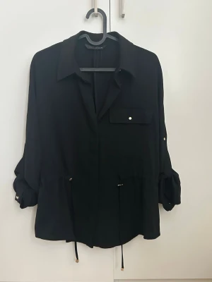 Snygg skjortjacka med snörning, ficka med tillhörande byxor från Zara - Stilren svart skjortjacka från Zara med krage, snörning i midjan och en bröstficka med knapp med tillhörande byxor, sparsamt använt. Perfekt för en trendig och avslappnad look.