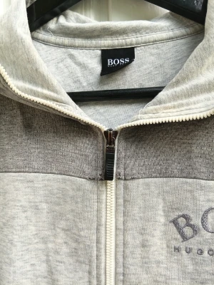 HUGO BOSS hoodie  - Snygg ljusgrå hoodie från HUGO BOSS med dragkedja framtill och logga på bröstet. Huvan har en dekorativ rand med BOSS-tryck. Tillverkad i mjukt bomullsmaterial och har en tvåtonad design med mörkare grå partier upptill.