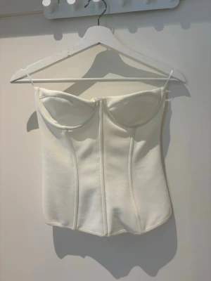 Vit bustier-body med tunna band - Säljer en stilren vit bustier-body med tunna axelband och markerade sömmar framtill. Bodyn har en figurnära passform och bygelkupor för extra stöd. Perfekt för att skapa en snygg siluett under olika outfits.