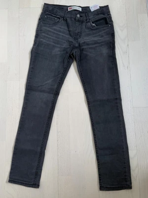 Mörkgrå Levis jeans - Snygga mörkgrå jeans med rak passform och klassisk femficksdesign. Jeansen har bälteshällor, knappgylf och subtila slitningar framtill för en avslappnad look. Perfekta till vardags och passar till det mesta i garderoben. Storlek W29 L30