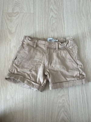 Beige shorts med fickor och uppvik - Snygga beige shorts med uppvikta benslut och praktiska sidofickor. Shortsen har hällor för bälte och två bakfickor. Perfekta för varma dagar och enkla att matcha med olika stilar. Tillverkade i mjukt bomullsmaterial för skön känsla.