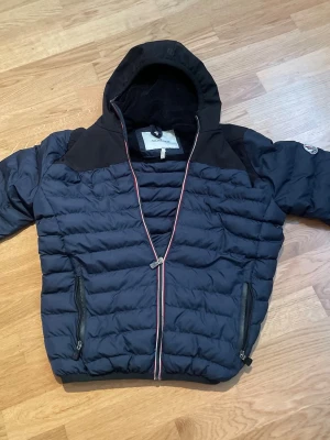 Mörkblå dunjacka med huva från Moncler - Snygg mörkblå dunjacka från Moncler med huva och dragkedja framtill. Jackan har två dragkedjeförsedda fickor och Moncler-logga på ärmen. Horisontella sömmar ger en puffig look och axelpartiet är i en matchande mörkare nyans. Perfekt för kalla vinterdagar.