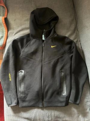 Svart Nike hoodie med dragkedja - Snygg svart hoodie från Nike med gul logga på bröstet och texten NOCTA på ärmen. Hoodien har hel dragkedja framtill, två fickor med dragkedja och en stor huva. Tillverkad i mjukt material som känns skönt mot huden. Perfekt för en sportig och avslappnad stil.