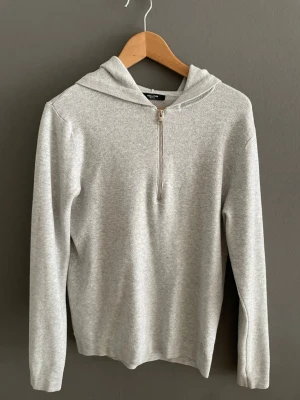 Ljusgrå Grisch hoodie med halv dragkedja - Snygg ljusgrå hoodie med huva och halv dragkedja framtill. Tillverkad i mjukt, stickat material som känns skönt mot huden. Långärmad modell med enkel och clean design, perfekt för en avslappnad look. Får en att se biffig ut.