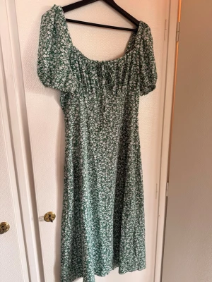 Grön blommig midiklänning - Söt grön klänning med vitt blommönster och puffärmar. Klänningen har en smickrande v-ringning och är i en luftig, flowy modell som passar perfekt till varma dagar. Materialet känns lätt och bekvämt, och den har en romantisk vintagekänsla.