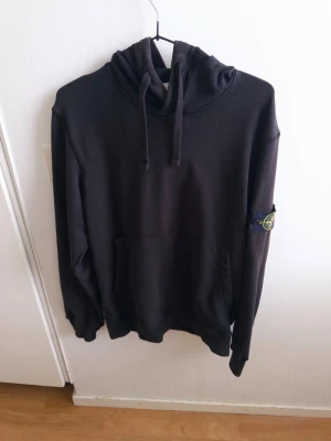 Svart hoodie från Stone Island - Säljer en svart hoodie från Stone Island med klassisk patch på vänster ärm. Hoodien har huva med dragsko och en stor magficka framtill. Tillverkad i mjukt bomullsmaterial och har ribbade muddar vid ärmslut och nederkant. Perfekt för en avslappnad streetwear-look.