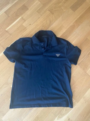 Svart pikétröja från Prada - Snygg marinblå pikétröja från Prada med klassisk krage och två knappar framtill. Tröjan har korta ärmar och den ikoniska Prada-loggan på bröstet. Perfekt för en clean och stilren look.