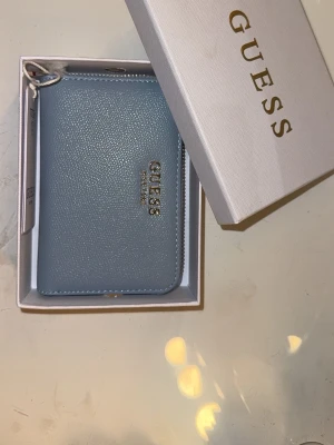 Ljusblå plånbok från Guess - Snygg ljusblå plånbok från Guess i ett stilrent och klassiskt format. Plånboken har strukturmönstrad yta och Guess-logga i guld framtill. Kommer i originalask, perfekt för dig som gillar trendiga accessoarer. Helt ny med prislapp, fick den i födelsedagspresent men använder tyvärr inte plånböcker🥰 kostade 700kr när den köptes därför säljs den för 600kr! Priset går att diskutera 🥰