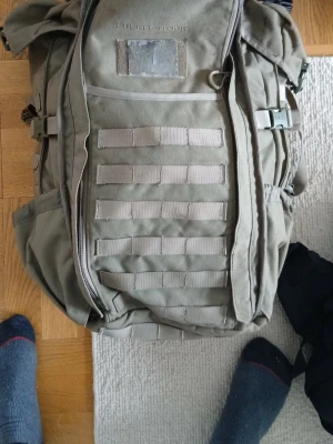 Taktisk ryggsäck i olivgrönt tyg - Rymlig taktisk ryggsäck i olivgrönt tyg med flera fack och MOLLE-system på framsidan. Justerbara axelremmar och sidofickor för extra förvaring. Perfekt för dig som gillar äventyr och vill ha en robust och funktionell väska.