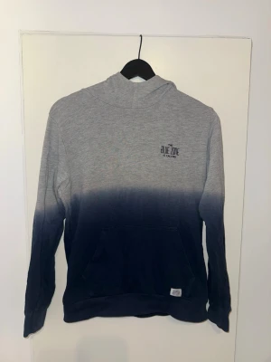 Grå och mörkblå hoodie med huva - Snygg hoodie i grått och mörkblått med cool dip dye-effekt. Tröjan har huva och en stor magficka framtill. Liten logga på bröstet där det står 'Blue Zone'. Perfekt för en avslappnad och trendig stil. Har en liten blå fläck på huvan men inget man tänker på. (Se bild).