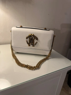 Vit handväska med guldkedja - Snygg vit handväska i rektangulär form med guldfärgad kedjerem och dekorativt guldfärgat monogram på framsidan. Väskan har stilrena linjer och är tillverkad i slätt material som ger en lyxig känsla. Perfekt accessoar för att lyfta din outfit.