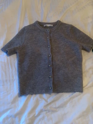 Grå kortärmad kofta med knappar - Kortärmad grå kofta med rund hals och ribbade kanter. Koftan har en rak passform och stängs med små knappar framtill. Materialet ser ut att vara mjukt och stickat, perfekt för lager på lager och en chill look.  Jättefin kofta ifrån Zara, använd 1 gång. Bra skick!!