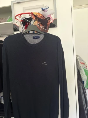 Mörkblå stickad tröja från GANT - Snygg mörkblå stickad tröja från GANT med rund halsringning och diskret logga broderad på bröstet. Tröjan har lång ärm och är tillverkad i ett mjukt, tunt stickat material som passar perfekt till lager på lager.